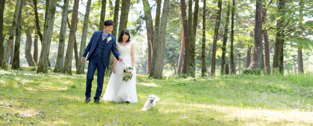 大分県別府市・別府公園の緑豊かな木立の中で、愛犬と一緒に散歩を楽しむ新郎新婦を撮影したフォトウェディングの一枚。