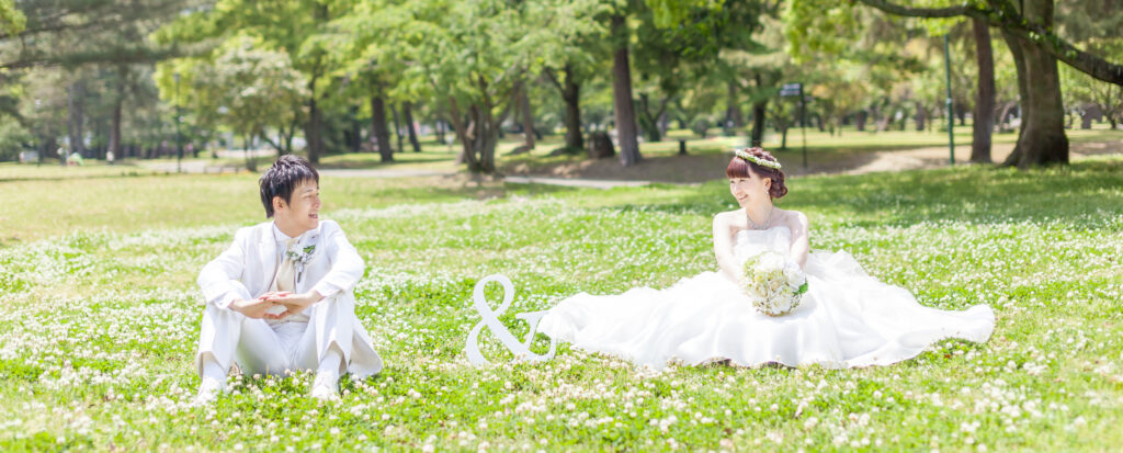 大分県別府市・別府公園のシロツメクサが広がる芝生で、笑顔で見つめ合う新郎新婦を撮影したフォトウェディングの一枚。