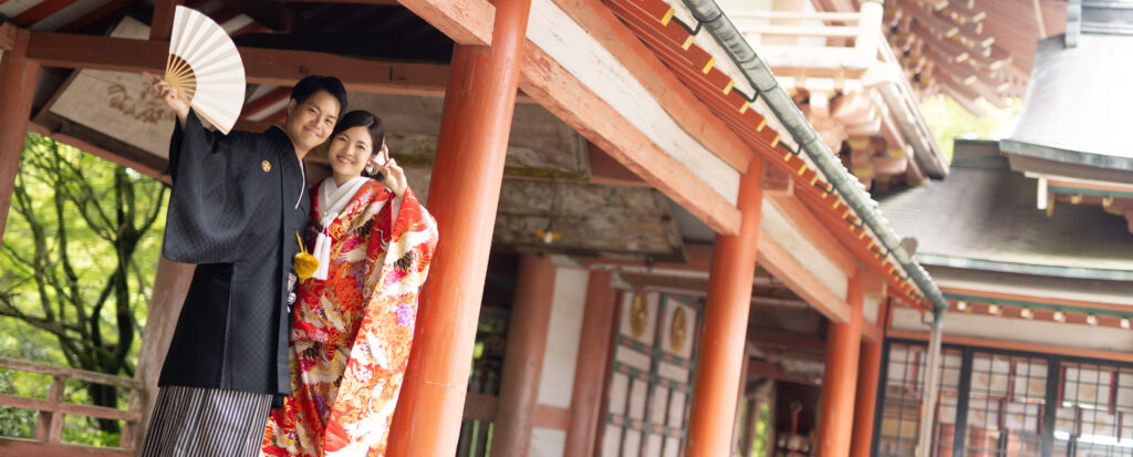 大分市の柞原八幡宮で和装前撮り。黒紋付の新郎と鮮やかな赤い色打掛の新婦が、歴史ある社殿を背景に笑顔を見せる姿。