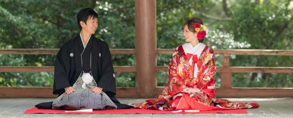 大分市の柞原八幡宮で和装前撮り。黒紋付袴の新郎と華やかな赤い色打掛の新婦が、緑に囲まれた回廊で向かい合い笑顔を交わす姿。