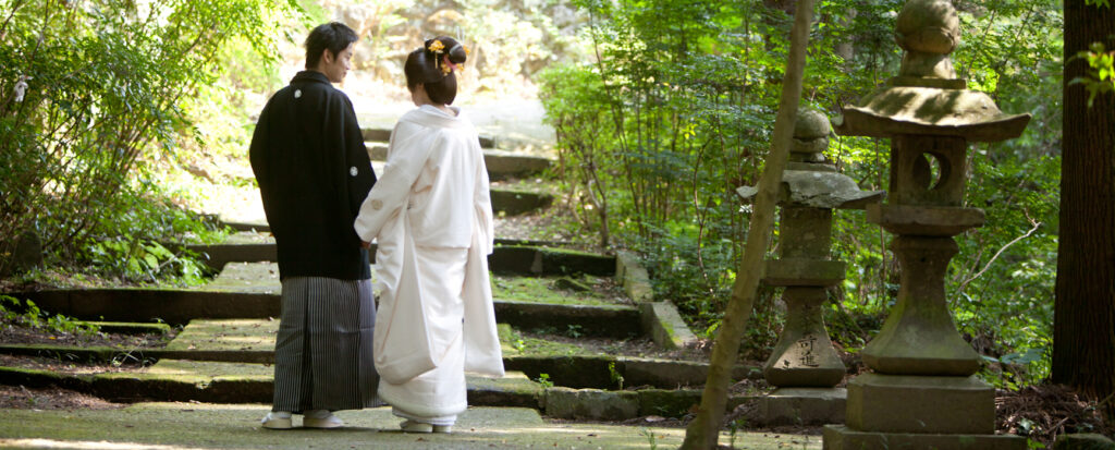 大分市の柞原八幡宮で和装前撮り。緑に囲まれた石段の参道で、黒紋付の新郎と白無垢の新婦が手をつなぎ寄り添う姿。