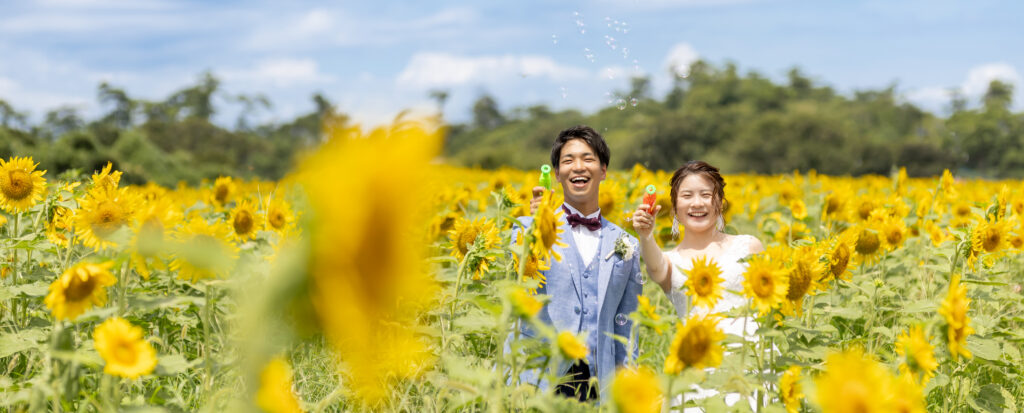 大分県豊後高田市の長崎鼻で洋装前撮り。青空の下、一面に咲くひまわり畑でシャボン玉を飛ばしながら笑顔を見せる新郎新婦。