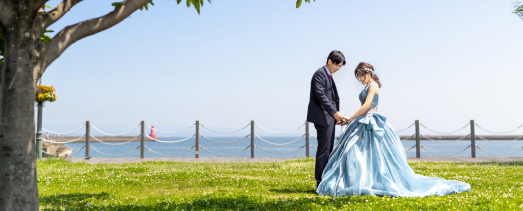 大分市・かんたん港園でフォトウェディング。海と緑の芝生を背景に、カラードレス姿で手を取り合う新郎新婦の一枚。