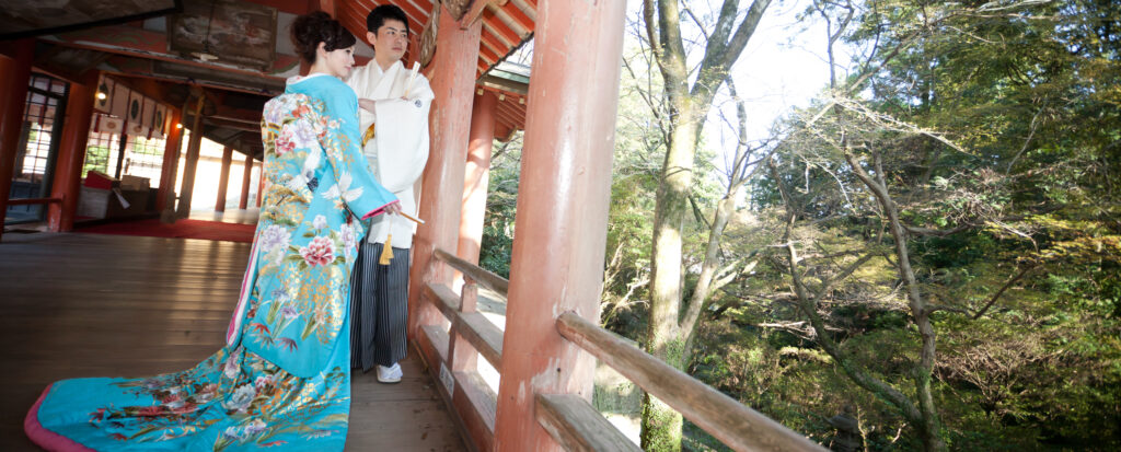 大分市の柞原八幡宮で和装前撮り。朱色の回廊で、鮮やかな水色の色打掛を着た新婦と白紋付の新郎が並び、境内の緑を眺める姿。