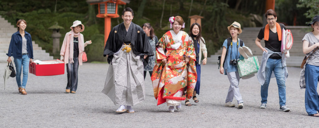 宇佐神宮での和装前撮りで、黒紋付袴の新郎と色打掛の新婦がスタッフと笑顔で歩くフォトウェディング