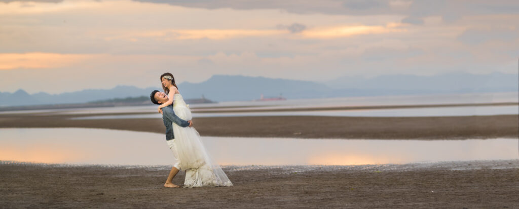 真玉海岸代替テキスト:
夕暮れの海辺で、ウエディングドレス姿の新婦を新郎が抱き上げて笑い合うフォトウェディング