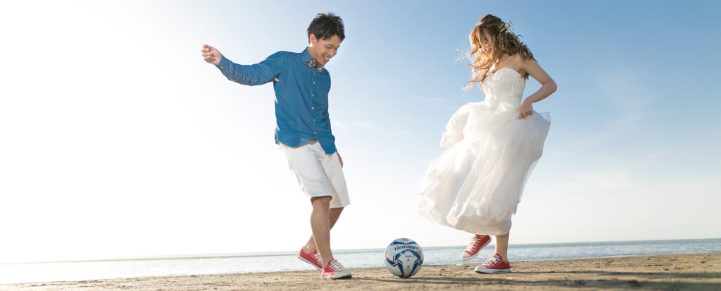 Photostudio PASSION青空が広がる海辺で、ウエディングドレス姿の新婦と新郎がサッカーボールで遊ぶフォトウェディング