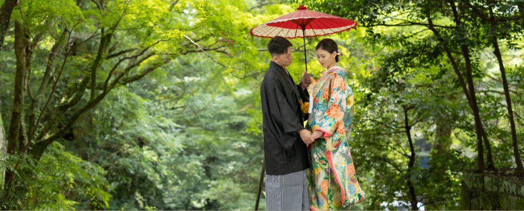 Photostudio PASSION鮮やかな新緑に囲まれたロケーションで、赤い和傘を差して寄り添う新郎新婦の和装フォトウェディング